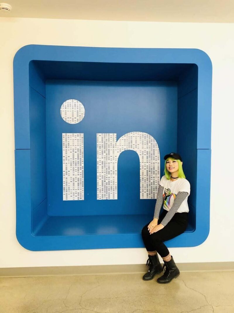 Goldie Chan, with green hair, sitting inside a large blue LinkedIn "in" logo sculpture.