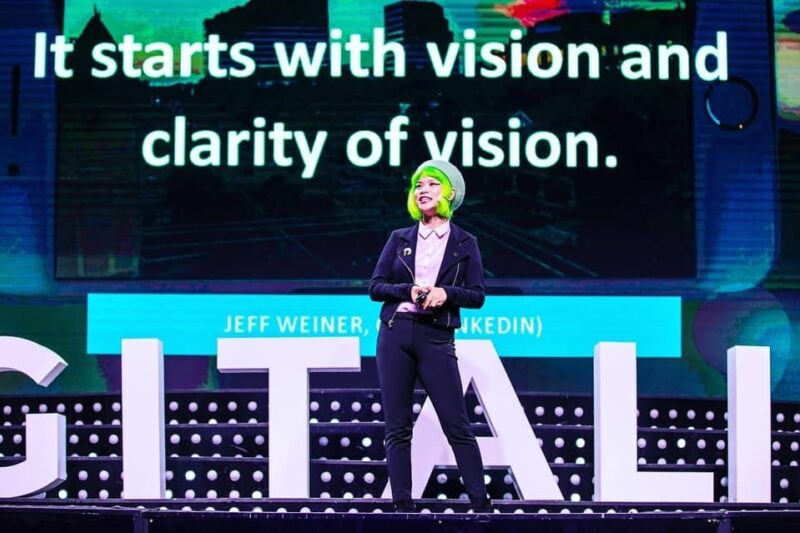 Goldie Chan, with bright green hair, standing on a conference stage in front of a large screen reading "It starts with vision and clarity of vision."